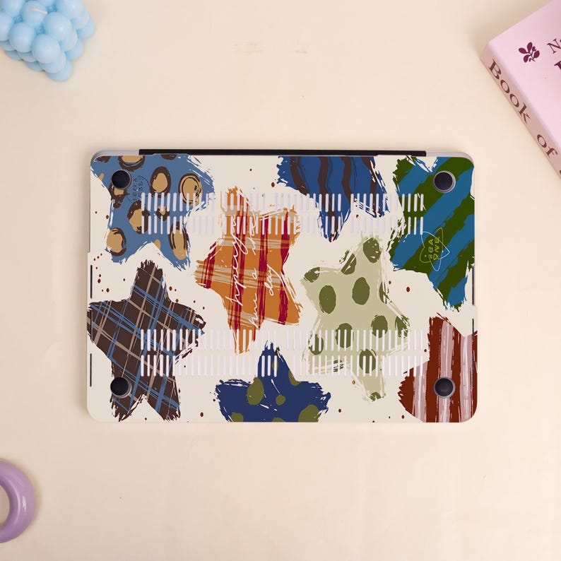 May include: A laptop case with a cream background and colorful star patterns. The stars feature various designs, including plaid, polka dots, and stripes in shades of blue, brown, orange, and green. The case has a vent design.