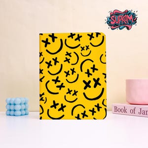 May include: A rectangular notebook with a bright yellow cover featuring a repeating pattern of black smiley faces with X-shaped eyes. The cover displays the text SUPKAM in a stylized, colorful graffiti-inspired font at the top right corner.
