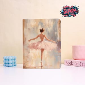 May include: A rectangular notebook featuring an illustration of a ballerina in a light pink tutu, standing on one leg against a soft, abstract beige and blue background. The top right corner displays the stylized text SUPKAM in blue and pink.