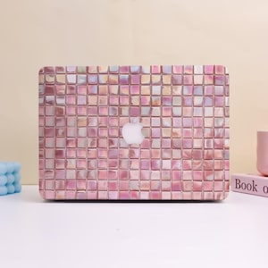 May include: A pink and iridescent laptop case with a mosaic tile pattern. The case features a white Apple logo in the center. The case is designed to fit a laptop and provides a decorative and protective covering.