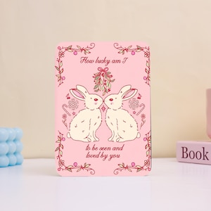 May include: A pink rectangular card with a decorative border and the text "How lucky am I" at the top. Two white rabbits face each other, with the text "to be seen and loved by you" below. The card has a festive design.