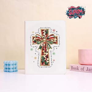Puede incluir: Un cuaderno blanco con un diseño de cruz con estampado de leopardo y un lazo verde y rojo. El texto "Jesus is the reason for the season" rodea la cruz. También se ven una vela de burbujas azul y una taza rosa.