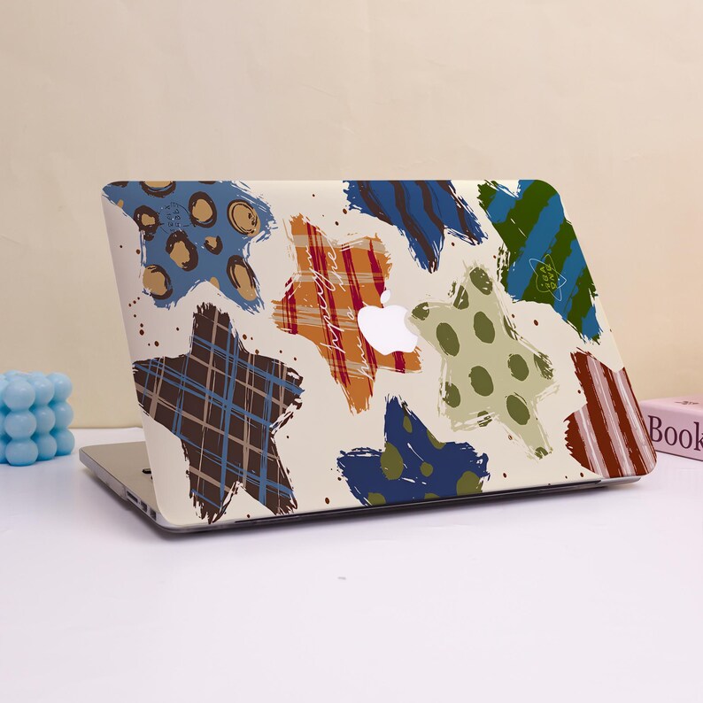 May include: Laptop computer with a cream-colored cover featuring colorful star-shaped designs. The stars have various patterns, including plaid, polka dots, and stripes in shades of blue, orange, and brown. The Apple logo is visible.