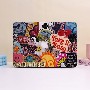 May include: A laptop case covered in colorful stickers. The stickers include a bridge, a heart, a star, cherries, and the words "Take It Easy" and "Darling". The case has a white Apple logo.