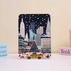 May include: A rectangular card featuring a winter scene of New York City. The illustration includes the Statue of Liberty, the Empire State Building, and a yellow taxi cab. Snowflakes fall against a dark blue sky, with a Christmas tree in the foreground.
