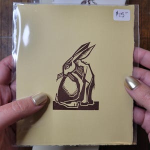 May include: A brown linocut print of a rabbit on a cream-colored paper. The rabbit is depicted in a stylized, geometric form. The print is in a clear plastic sleeve. The price tag reads $15.