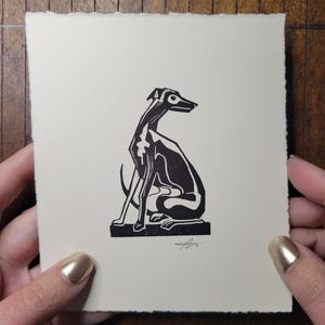 May include: A black and white linocut print of a seated greyhound dog. The artwork is on a cream-colored paper with a deckled edge. The dog is depicted in a stylized, minimalist design.