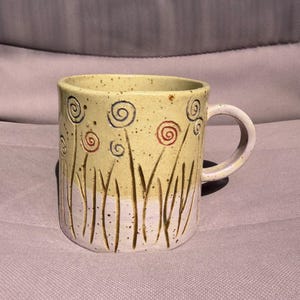 Pode incluir: Uma caneca de cerâmica com acabamento salpicado, apresentando um design bicolor de amarelo pálido na metade superior e branco na inferior. A superfície é decorada com linhas verticais esculpidas e padrões de espirais em azul e laranja.