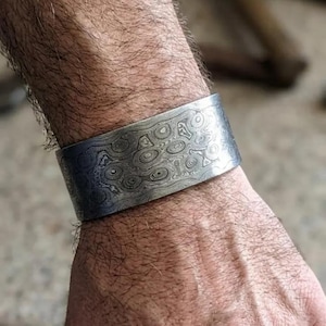 Puede incluir: Un brazalete de metal ancho y estampado con un diseño en espiral. El brazalete plateado se lleva en la muñeca, mostrando el intrincado patrón. El brazalete mide aproximadamente 5 cm de ancho.
