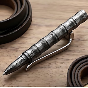 Puede incluir: Un bolígrafo plateado y negro con un diseño similar al bambú, con un clip. El bolígrafo está sobre una superficie de madera, con un cinturón de cuero en el fondo. El cuerpo del bolígrafo tiene una apariencia texturizada de vetas de madera.