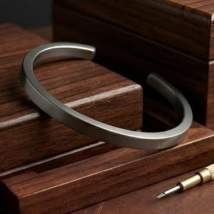 May include: A silver cuff bracelet with a flat, rectangular design. The bracelet is open at the bottom and rests on a dark wooden surface. A small screwdriver is also visible, suggesting jewelry repair or adjustment.