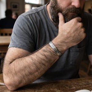 Könnte beinhalten: Ein silbernes Armband mit geometrischem Design ist am Arm zu sehen. Die Person trägt ein graues T-Shirt und sitzt an einem Holztisch. Ein offenes Buch liegt auf dem Tisch. Die Szene spielt in einem Café oder Restaurant.