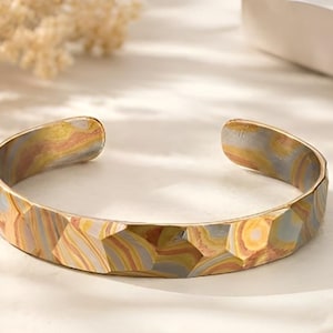 Puede incluir: Un brazalete de puño texturizado con un patrón geométrico. El brazalete abierto presenta una mezcla de colores, incluyendo dorado, gris y marrón. Esta joya está diseñada para ser usada en la muñeca.