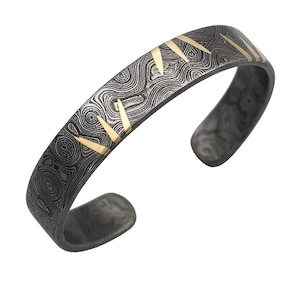 Pode incluir: Uma pulseira de punho cinza escuro com uma superfície estampada e detalhes triangulares dourados. A pulseira aberta tem um design texturizado e rodopiante. Os detalhes dourados são espaçados na parte superior.