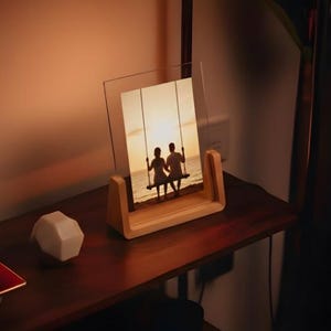 Pode incluir: Uma moldura de madeira com uma frente de vidro transparente exibe uma cena de pôr do sol de duas pessoas em um balanço. A moldura tem um acabamento de madeira natural e fica em uma prateleira de madeira escura. Um objeto geométrico branco e uma pequena bandeja vermelha também estão na prateleira.