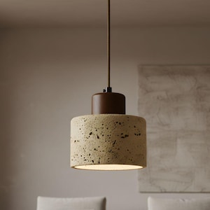 May include: A contemporary pendant light featuring a cylindrical, textured beige shade and a dark brown top. It hangs from a brown cord, providing a warm, ambient glow. The light is designed for indoor use.