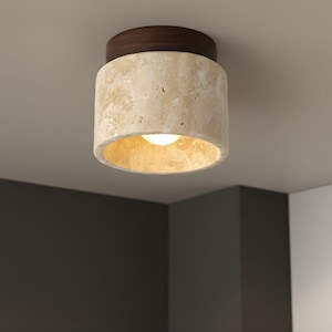 Może przedstawiać: Cylindryczna lampa sufitowa z teksturowanym, beżowym korpusem przypominającym kamień i ciemnobrązowym drewnianym blatem. Światło ma ciepłe, złote wnętrze i widoczną żarówkę. Oprawa jest zamontowana na białym suficie.