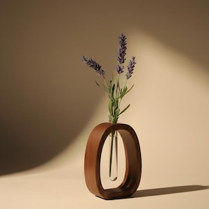 Op de afbeelding: Een houten vaas met een ovaal frame bevat een heldere glazen buis met lavendel takjes. De vaas is gemaakt van donkerbruin hout en staat op een lichtbeige oppervlak. De lavendel heeft paarse bloemen en groene stelen.