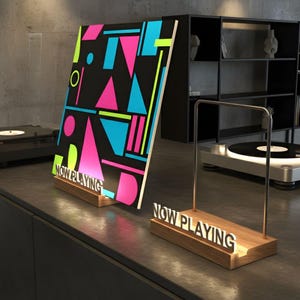 Puede incluir: Soporte de exhibición de discos de vinilo de madera con las palabras "NOW PLAYING" en blanco. El soporte sostiene un disco de vinilo con un diseño geométrico colorido en rosa, azul y verde. También se ve un segundo soporte.