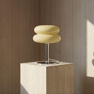 Puede incluir: Una lámpara de mesa de estilo retro con una pantalla apilada y redondeada en un tono amarillo suave. La lámpara presenta un vástago y una base cromados, y se asienta sobre un pedestal cuadrado de color claro. El diseño evoca una estética moderna de mediados de siglo.