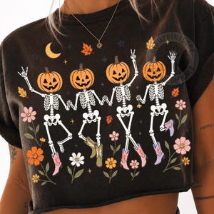 May include: A black short-sleeve t-shirt featuring a graphic of four skeletons with pumpkin heads wearing cowboy boots. The skeletons are dancing among colorful flowers, autumn leaves, and a crescent moon. The design includes small stars.