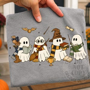 May include: A heather gray t-shirt featuring a graphic print of four white ghosts dressed in scarves and accessories. The ghosts are depicted with items including a broom, a wizard hat, books, a golden snitch, and a wand, all in a cartoon style.