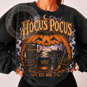 May include: A charcoal black crewneck sweatshirt featuring a large graphic print of a jack-o'-lantern, a bubbling cauldron, a black cat, a broom, and books labeled Spells, Potions, Brews, and Book of Shadows with the text Hocus Pocus and Salem.