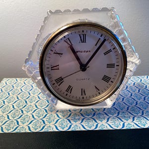May include: A clear, faceted quartz clock with a silver face and black Roman numerals. The clock face has the word "STAGER" and "QUARTZ" printed on it. The clock is set in a hexagonal, clear, decorative frame.