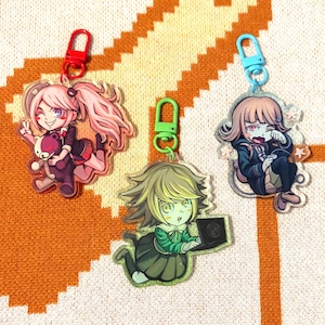 Puede incluir: Tres llaveros acrílicos de personajes de anime con cierres coloridos. Los personajes están en varias poses, uno de ellos sosteniendo un oso de peluche. Los llaveros están sobre una superficie naranja y blanca texturizada.