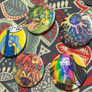 Può includere: Una collezione di cinque bottoni rotondi e colorati con varie illustrazioni in stile anime. I bottoni presentano accenti olografici e design vivaci, tra cui personaggi ed elementi grafici. I bottoni sono esposti su una superficie a motivi.