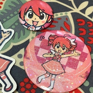 Può includere: Una collezione di bottoni e un adesivo di personaggi in stile anime rosa e rossi. I bottoni raffigurano una ragazza sorridente con i capelli rossi e un vestito rosa. Un bottone è piccolo, l'altro è più grande con uno sfondo a scacchi. L'adesivo è un ritaglio dello stesso personaggio.
