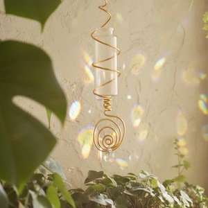 May include: A decorative hanging glass vial with a gold-colored wire frame. The frame features spiral and wavy designs. The vial is suspended in front of a textured wall, with plants in the foreground.