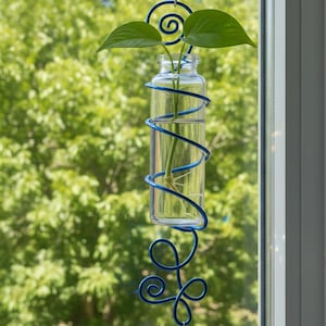 May include: A clear glass bottle with a blue wire wrap, holding a plant with green leaves. The bottle hangs from a suction cup on a window. A decorative glass bead hangs below.