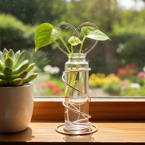 May include: A clear glass vase with a gold-colored wire heart-shaped frame holding green-leafed plants. The vase sits on a wooden surface near a window, with a succulent in a white pot to the left. The background shows a blurred garden.