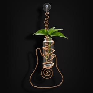 May include: A copper wire guitar-shaped vase with a clear glass bottle holding green leafy plants. The vase is attached to a black wall with a suction cup. The copper wire is coiled around the bottle and forms the guitar's outline.
