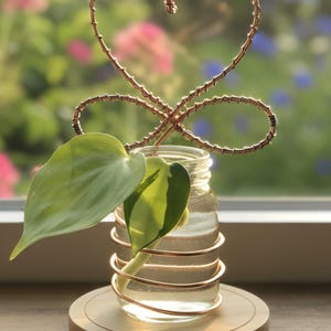 May include: A small glass jar with a copper wire design holding a plant with green leaves. The jar sits on a round wooden base. A copper wire heart shape is above the jar. The background is a blurred view of a garden.