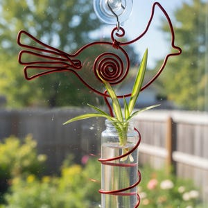 May include: A red wire bird-shaped plant holder with a glass jar containing a green plant. The jar is wrapped with red wire and hangs from a suction cup on a window. The background shows a blurred outdoor scene.