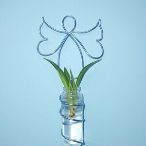 May include: A blue wire angel-shaped wall vase holding a small glass bottle with water and green leaves. The wire angel has outstretched wings and a halo. The bottle is wrapped in wire.