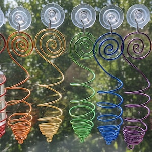 May include: A row of colorful, handcrafted wire plant holders with glass vials, attached to a window with suction cups. Each holder is a different color of the rainbow, with a spiral design and a small glass bottle to hold a plant cutting.