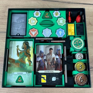 Può includere: Un vassoio organizzatore per giochi da tavolo contenente vari componenti, tra cui mazzi di carte con i personaggi Nisaba e Schwindel, gettoni verdi con simboli, cubi gialli e neri e monete metalliche circolari contrassegnate con 1, 3 e 6.
