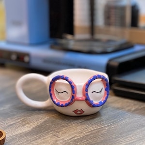 Può includere: Una tazza di ceramica bianca con manico nero e la scritta "But First, Coffee" in grassetto nero. La tazza è posta su uno sfondo sfocato, che suggerisce un'atmosfera accogliente.