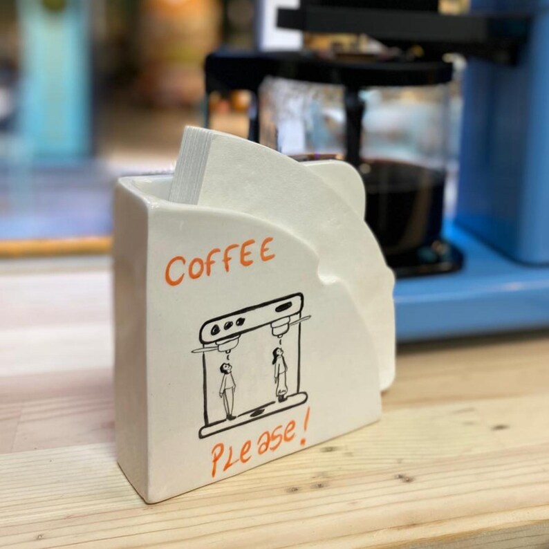 Może przedstawiać: Biały ceramiczny uchwyt na filtr do kawy ze słowem "Coffee" w pomarańczowym skrypcie i frazą "Please!" w pomarańczowym skrypcie. Na uchwycie znajduje się r&oacute;wnież czarno-biała ilustracja ekspresu do kawy. Uchwyt jest wypełniony białymi filtrami.