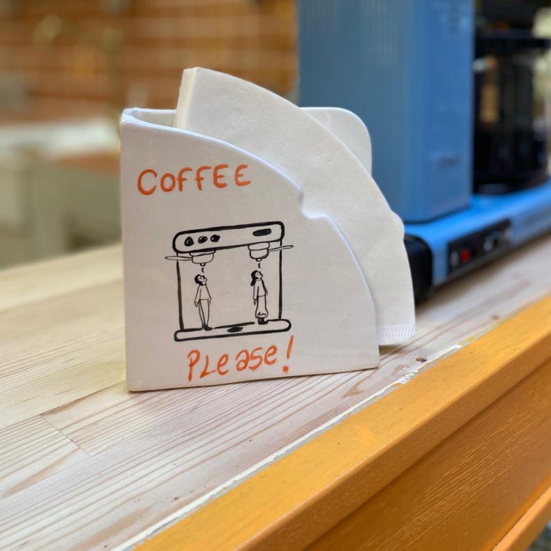 May include: White ceramic coffee filter holder with the word "Coffee" in orange and the phrase "Please!" in black. The holder has a black illustration of two figures under a coffee machine. White coffee filters are inside.