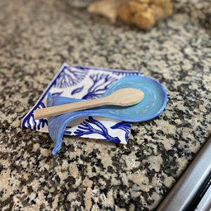May include: A blue ceramic spoon rest with a matching holder, resting on a white cloth with blue coral designs. A wooden spoon rests in the spoon rest. The kitchenware is on a speckled countertop.