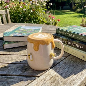 May include: A white ceramic mug with a honey-like glaze and two bee embellishments. The mug is filled with a latte, and rests on a wooden table next to books with the titles "Garden Wisdom" and "The Hidden Life of Trees".