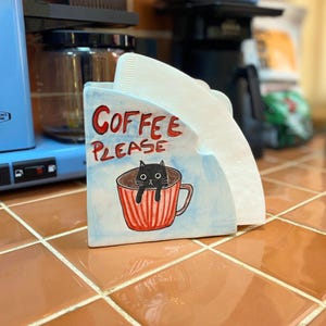 May include: A ceramic coffee filter holder with a light blue background and a hand-painted illustration of a black cat inside a red and white striped mug. The text COFFEE PLEASE is written in red above the cat. White paper filters are inside.