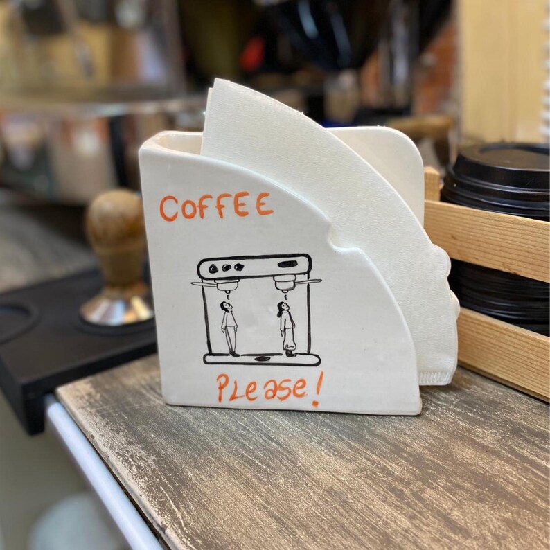 Może przedstawiać: Biały ceramiczny uchwyt na filtr do kawy ze słowem "COFFEE" w kolorze pomarańczowym i czarno-białą ilustracją dw&oacute;ch postaci pod ekspresem do kawy. Poniżej ilustracji znajdują się słowa "Please!". Uchwyt zawiera białe filtry papierowe.