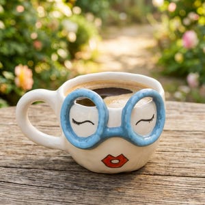 May include: A white ceramic mug featuring a hand-painted face with closed eyes, a pair of raised light blue glasses, and red lips. The mug is filled with dark coffee and has a rounded handle on the left side, showcasing a smooth, glossy finish.