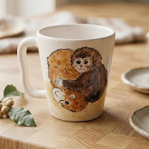 May include: A white ceramic mug with a curved handle. The mug features a watercolor illustration of a brown monkey hugging a smaller, light brown animal. The background is a light yellow. The mug is on a wooden surface.