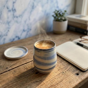 Op de afbeelding: Een keramische koffiemok met blauw-witte strepen, gevuld met koffie, staat op een houten tafel. Een bijpassende schotel staat links. Stoom stijgt op uit de koffie. Een notitieboek, pen en kleine plant staan ook op de tafel.